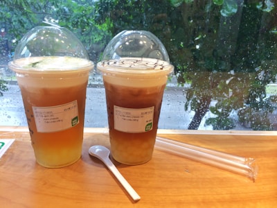 Two cups of iced tea with cream cheese topping are placed on a wooden table. The drinks have dome-shaped lids and are labeled with a sticker. Next to the cups is a wrapped straw and a plastic spoon. Raindrops are visible on the window behind, with a blurred view of greenery outside.