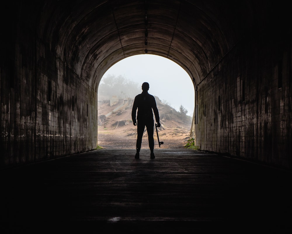 silhouette photo of a man carrying skateboard inside tunnel