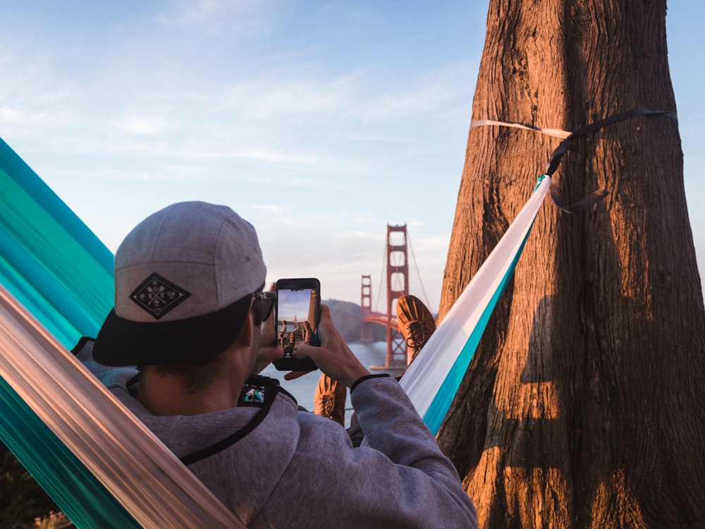 shallow focus photography of man on hammock while taking photo of Golden Gate Bridge of California to his phone
