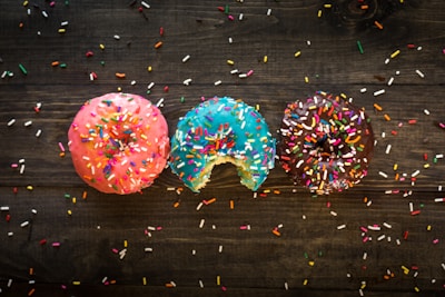 Three donuts with colorful icing are sprinkled with various rainbow sprinkles. The donuts are pink, blue, and brown in color, and one donut has a bite taken out of it. They rest on a dark wooden surface scattered with additional sprinkles.