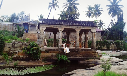An ancient stone structure with pillars stands in a scenic environment surrounded by lush greenery and tall palm trees. The background features an old temple building adorned with intricate carvings. A person dressed in traditional attire is seated on the steps of the structure, adding a cultural and historical essence to the scene.