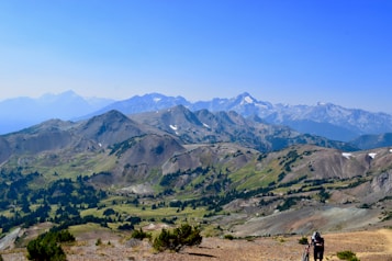 A panoramic view of rugged mountain ranges with patches of vegetation and snow atop some peaks. The horizon showcases a clear blue sky above the distant mountains. In the foreground, a person wearing outdoor gear is seen carrying a bicycle uphill, adding a sense of adventure.
