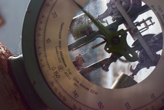 A close-up view of an old mechanical scale with a round dial and Cyrillic writing. The intricate gears and levers are visible through the glass cover, indicating its complex mechanism. A rustic appearance with evident wear and a slightly dusty surface suggests age and usage.