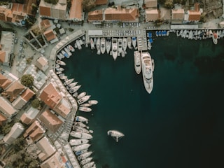 An aerial view of a marina with numerous boats and yachts docked. The marina is adjacent to a coastal town with buildings featuring red-tiled roofs. The turquoise water of the marina contrasts with the earthy tones of the buildings and concrete elements of the harbor. A single boat is floating near the center of the marina space.