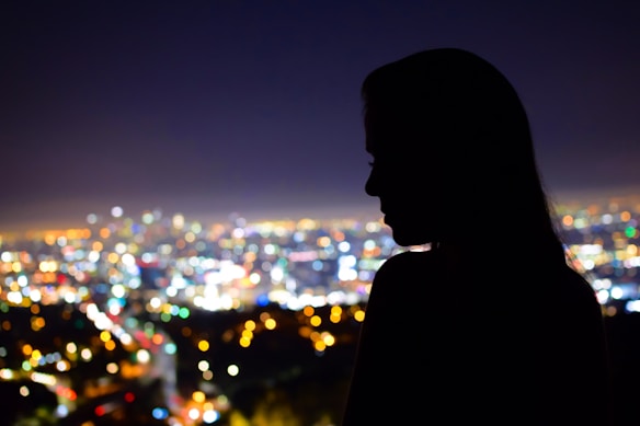 A silhouette of a person is visible against a backdrop of a vibrant cityscape at night. The city lights twinkle in various colors, creating a bokeh effect that blurs the details of the buildings below. The horizon is slightly illuminated, suggesting an urban environment.
