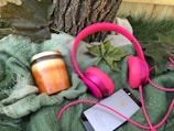 A set of vibrant pink headphones, a jar candle with layers of orange and yellow, and a smartphone with a reflective surface rest on a textured green blanket. Large green leaves and a tree trunk add a touch of nature to the scene.