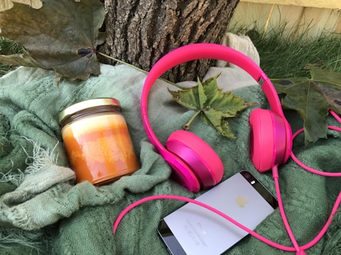 A set of vibrant pink headphones, a jar candle with layers of orange and yellow, and a smartphone with a reflective surface rest on a textured green blanket. Large green leaves and a tree trunk add a touch of nature to the scene.