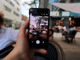 A hand holds a smartphone capturing an image of a bustling outdoor cafe. The foreground shows the phone displaying the scene, while the background is blurred, depicting a lively urban environment with people sitting at tables under an outdoor canopy.