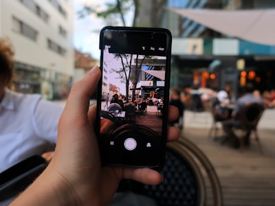 A hand holds a smartphone capturing an image of a bustling outdoor cafe. The foreground shows the phone displaying the scene, while the background is blurred, depicting a lively urban environment with people sitting at tables under an outdoor canopy.