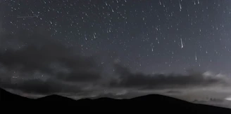 A night sky illuminated by a bright shooting star streaking over rolling hills.