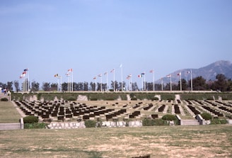 A serene cemetery with rows of identical headstones is lined up across a well-maintained lawn. Numerous flagpoles with different national flags are visible in the background, suggesting a memorial setting. Trees and mountains form a distant backdrop under a clear blue sky.