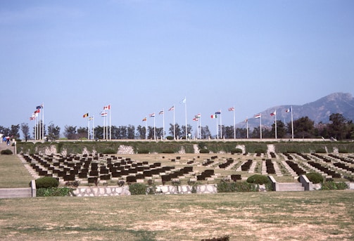 A serene cemetery with rows of identical headstones is lined up across a well-maintained lawn. Numerous flagpoles with different national flags are visible in the background, suggesting a memorial setting. Trees and mountains form a distant backdrop under a clear blue sky.