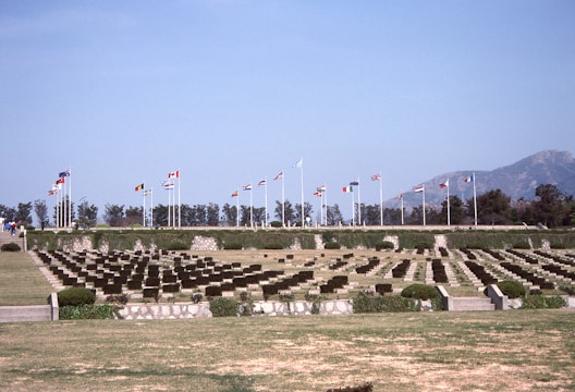 A serene cemetery with rows of identical headstones is lined up across a well-maintained lawn. Numerous flagpoles with different national flags are visible in the background, suggesting a memorial setting. Trees and mountains form a distant backdrop under a clear blue sky.