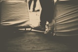 Sepia-toned photograph of a person's legs and feet extended, with the person seated on a covered chair. The background appears blurred. The image focuses on the feet and lower legs, with the shoes tied tightly.