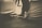 Sepia-toned photograph of a person's legs and feet extended, with the person seated on a covered chair. The background appears blurred. The image focuses on the feet and lower legs, with the shoes tied tightly.