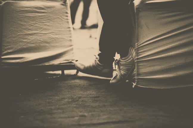Sepia-toned photograph of a person's legs and feet extended, with the person seated on a covered chair. The background appears blurred. The image focuses on the feet and lower legs, with the shoes tied tightly.