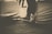 Sepia-toned photograph of a person's legs and feet extended, with the person seated on a covered chair. The background appears blurred. The image focuses on the feet and lower legs, with the shoes tied tightly.