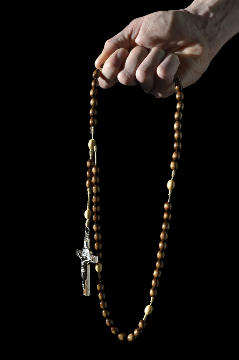 A hand gently holds a wooden rosary with a crucifix pendant. The beads are made of dark wood, interspersed with lighter beads, connected by a thin string. The background is black, highlighting the rosary and the hand.