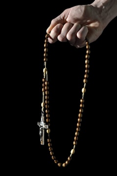 A hand gently holds a wooden rosary with a crucifix pendant. The beads are made of dark wood, interspersed with lighter beads, connected by a thin string. The background is black, highlighting the rosary and the hand.