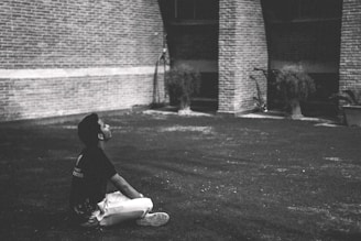 A person sits cross-legged on a grassy area, gazing upwards thoughtfully. The background features brick columns and some potted plants, creating an urban, introspective setting.