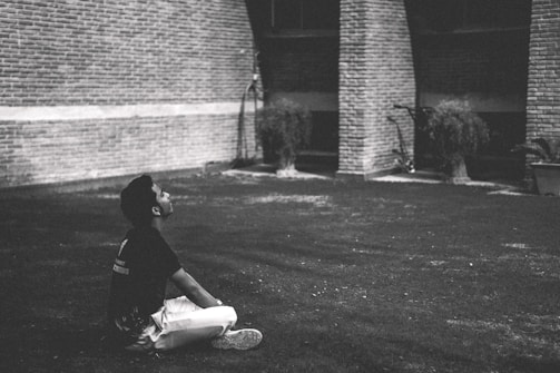 A person sits cross-legged on a grassy area, gazing upwards thoughtfully. The background features brick columns and some potted plants, creating an urban, introspective setting.