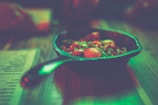 A skillet containing colorful vegetables such as cherry tomatoes and herbs sits on a wooden table. The lighting casts a greenish glow over the scene, giving the image an atmospheric and moody feel. The edges of a menu or paper are visible near the skillet.