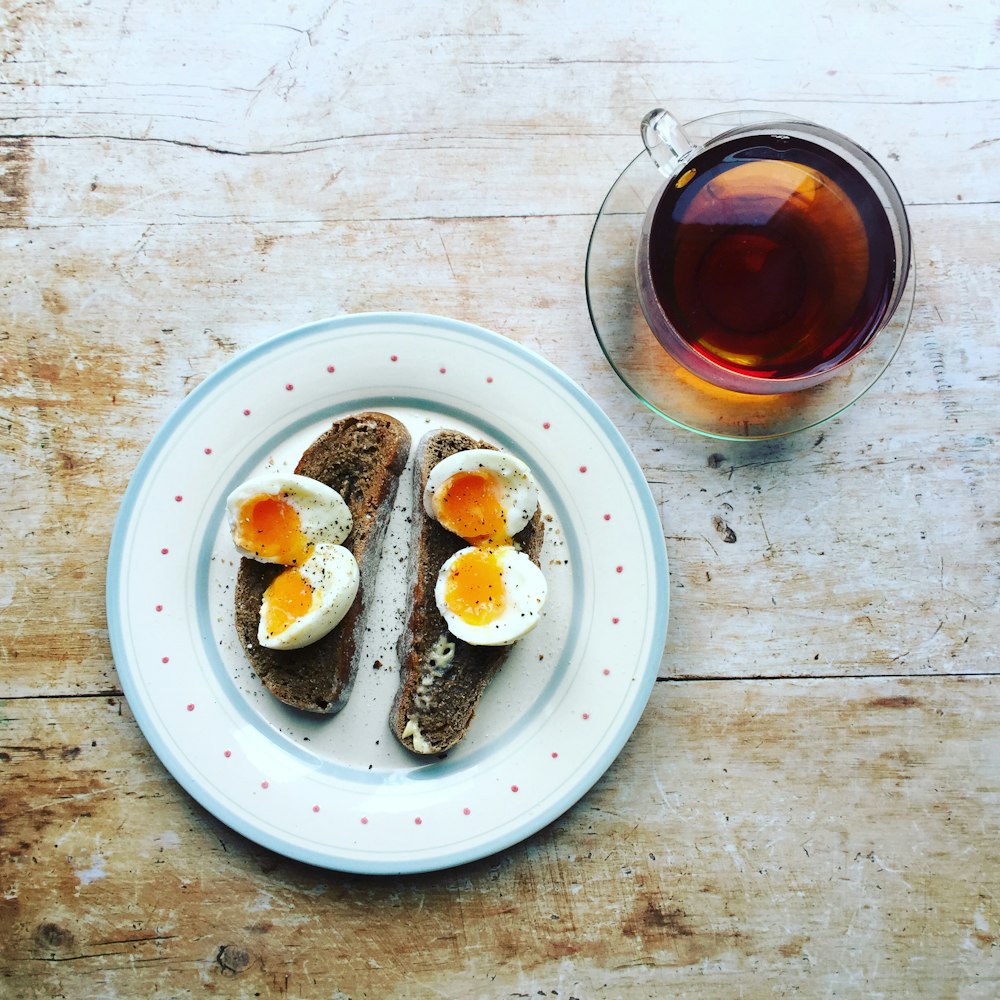 boiled eggs and toast on white ceramic plate near clear glass teacup filled with brown liquid