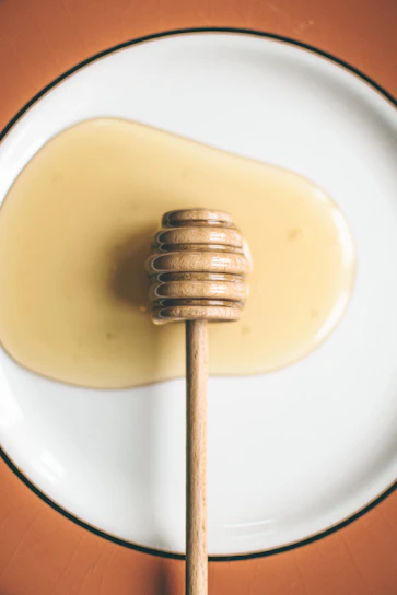 Close-up of golden macadamia honey dripping from a wooden honey dipper with macadamia tree blossoms in the background.