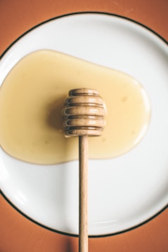 A wooden honey dipper rests on a white plate, drizzled with golden honey that spreads out around it. The background features a warm, soft orange hue creating an inviting, appetizing scene.