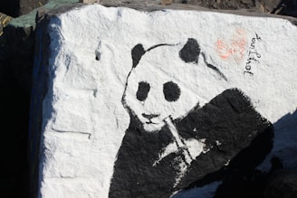 A black and white painting of a panda on a large rock. The panda is depicted munching on bamboo, and the left side of the image features textured stone surface with white and black paint. There is a small signature or writing near the top right corner of the panda.