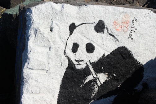 A black and white painting of a panda on a large rock. The panda is depicted munching on bamboo, and the left side of the image features textured stone surface with white and black paint. There is a small signature or writing near the top right corner of the panda.
