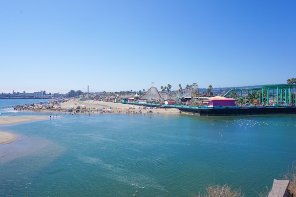 A lively beach scene with people enjoying the sand and ocean. An amusement park with roller coasters and colorful rides is visible in the background, adding a sense of fun and excitement. Palm trees line the area, and the clear blue sky enhances the summery atmosphere.