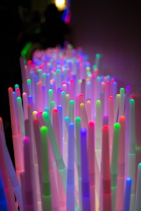 An array of upright, colorful LED light sticks, each glowing with vibrant hues of red, green, blue, purple, and pink, creating a mesmerizing visual effect in a dimly lit setting.