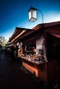A vibrant market stall displaying a variety of goods, including candies and snacks, under a blue sky. The stall is shaded by a prominent structure with a lantern-style light fixture above.
