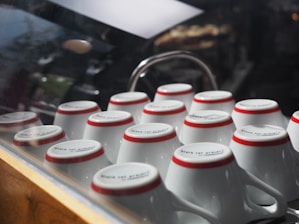 A collection of upside-down white ceramic cups with red rims arranged neatly on a countertop, reflecting light through a glass barrier.