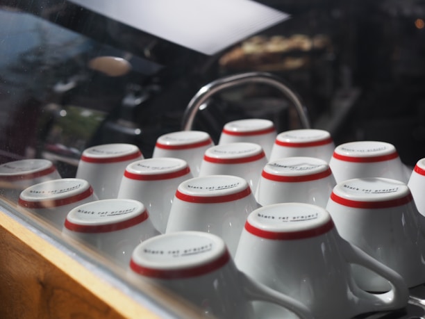 A collection of upside-down white ceramic cups with red rims arranged neatly on a countertop, reflecting light through a glass barrier.