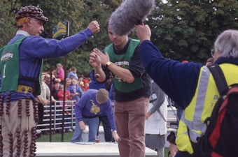 A group of people are gathered outdoors at an event, with two individuals engaged in a competition. One person is wearing a colorful costume adorned with chestnuts and a helmet-like hat, while the other person is holding a string with one chestnut attached. The competition seems to involve striking the chestnut with another. Spectators are visible in the background, and a person is holding a large microphone, likely indicating media coverage.