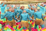 A group of dancers dressed in vibrant blue costumes adorned with metallic and leaf-like accessories perform under bamboo poles. They wear colorful skirts with layers of pink, green, and yellow ruffles. The dancers are synchronized, leaning backward while balancing and holding hands, creating a lively and dynamic scene.