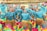 A group of dancers dressed in vibrant blue costumes adorned with metallic and leaf-like accessories perform under bamboo poles. They wear colorful skirts with layers of pink, green, and yellow ruffles. The dancers are synchronized, leaning backward while balancing and holding hands, creating a lively and dynamic scene.