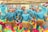 A group of dancers dressed in vibrant blue costumes adorned with metallic and leaf-like accessories perform under bamboo poles. They wear colorful skirts with layers of pink, green, and yellow ruffles. The dancers are synchronized, leaning backward while balancing and holding hands, creating a lively and dynamic scene.