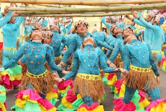A group of dancers dressed in vibrant blue costumes adorned with metallic and leaf-like accessories perform under bamboo poles. They wear colorful skirts with layers of pink, green, and yellow ruffles. The dancers are synchronized, leaning backward while balancing and holding hands, creating a lively and dynamic scene.