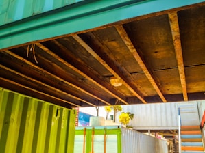 A view of the underside of a structure made of shipping containers. The containers are painted green and blue, and there are visible rusty beams supporting the structure. There is a light fixture mounted on the beams. In the background, a set of stairs is visible along with some trees and parts of another container.