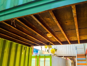A view of the underside of a structure made of shipping containers. The containers are painted green and blue, and there are visible rusty beams supporting the structure. There is a light fixture mounted on the beams. In the background, a set of stairs is visible along with some trees and parts of another container.