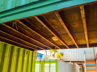 A view of the underside of a structure made of shipping containers. The containers are painted green and blue, and there are visible rusty beams supporting the structure. There is a light fixture mounted on the beams. In the background, a set of stairs is visible along with some trees and parts of another container.