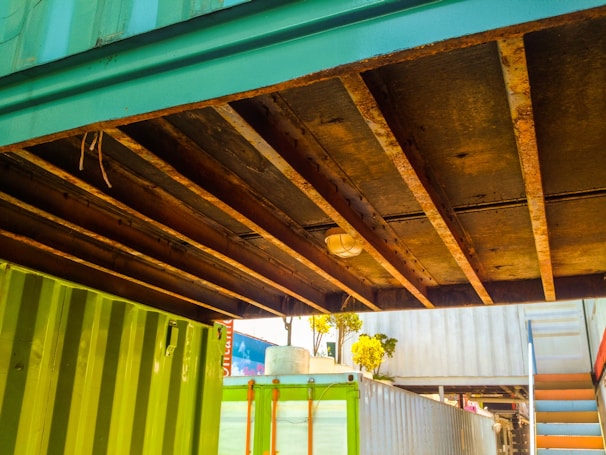 A view of the underside of a structure made of shipping containers. The containers are painted green and blue, and there are visible rusty beams supporting the structure. There is a light fixture mounted on the beams. In the background, a set of stairs is visible along with some trees and parts of another container.