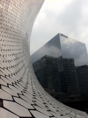 A unique architectural structure featuring a curved facade made of hexagonal tiles is prominently displayed. In the background, there is a modern glass-clad building with reflective surfaces. The image gives a sense of contemporary urban design, merging art with architecture.