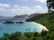 A picturesque tropical beach surrounded by lush greenery and clear turquoise water. A line of palm trees extends along the white sandy shore. In the distance, small islands are visible near the tranquil sea. Fluffy clouds float in the sky, creating a peaceful and serene atmosphere.