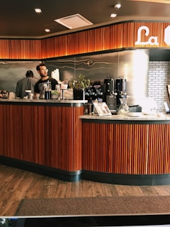 A warmly lit coffee shop interior with a modern design. The counter is made of wood with vertical slats, giving it an elegant appearance. There are two people behind the counter, one interacting with something on the counter and the other appearing to wait or observe. Various coffee-making equipment is visible, including espresso machines and a coffee grinder. The backdrop features a wave-like design on the wall and part of the logo of the coffee shop is visible at the top right.