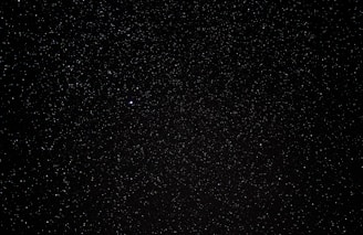 A vast expanse of a night sky filled with countless tiny stars scattered across a deep black background, with a single noticeably brighter star. The scene embodies the vastness of the universe.
