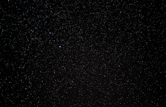 A vast expanse of a night sky filled with countless tiny stars scattered across a deep black background, with a single noticeably brighter star. The scene embodies the vastness of the universe.